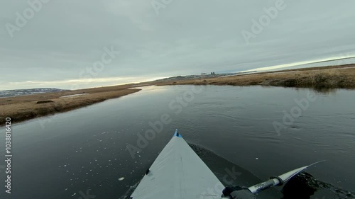 A point of view shot from a kayak as it moves towards a town. The kayak is moving through cold water with small pieces of ice floating in it.