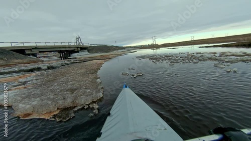 First-person view kayak paddling through icy waters towards a bridge on a cloudy day.