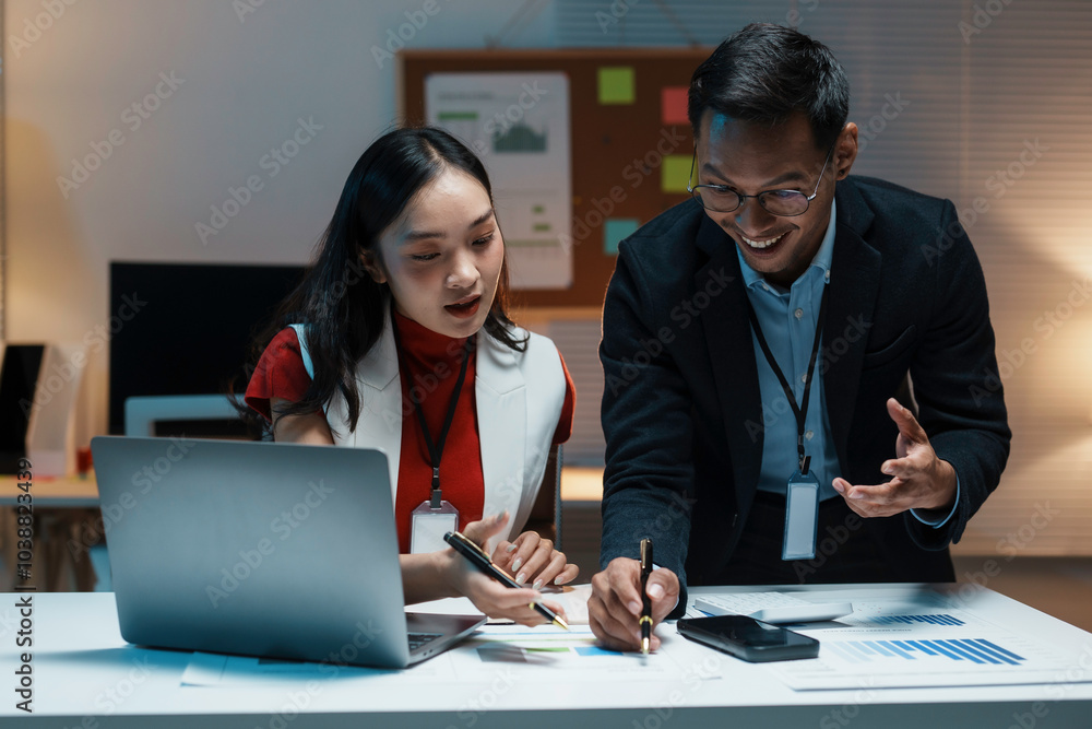 Two young professionals discussing a project, looking at charts and using a laptop, working late in the office Two young professionals discussing a project, looking at charts and using a laptop, working late in the office