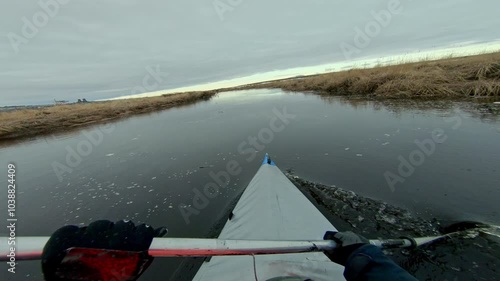 A solo kayaker paddles through a narrow creek, surrounded by a landscape of brown grasses and a cloudy sky