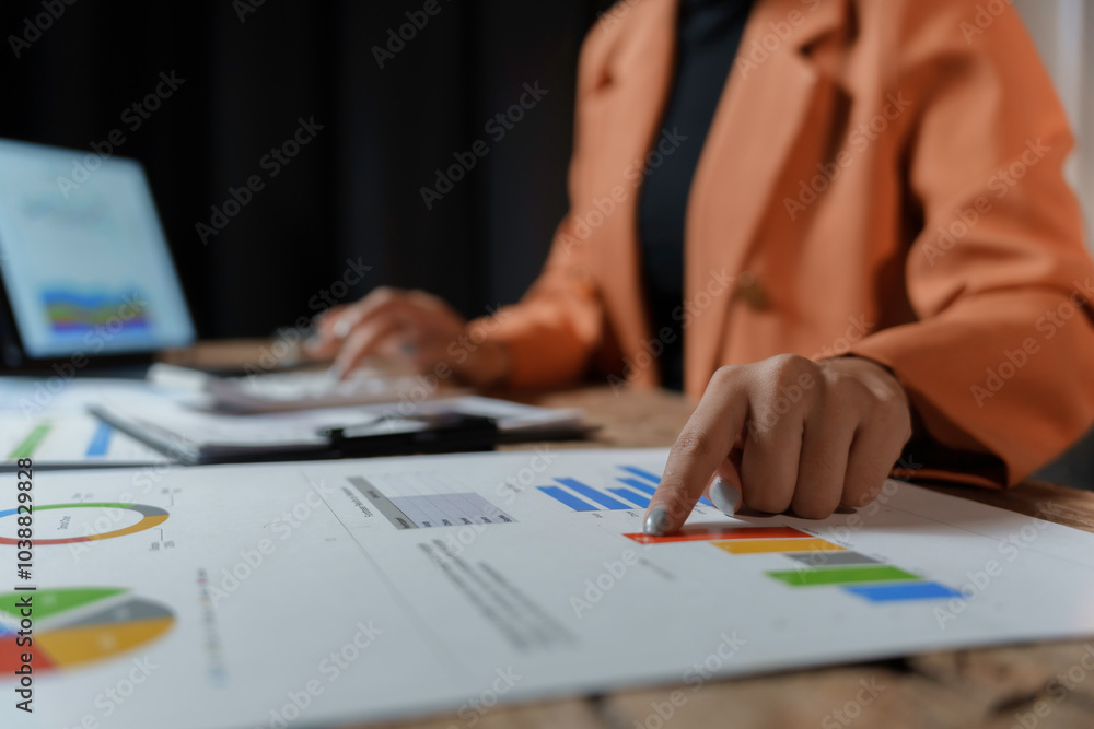 Businesswoman is working late, analyzing financial charts and using a calculator to review her company's performance Businesswoman is working late, analyzing financial charts and using a calculator to review her company's performance