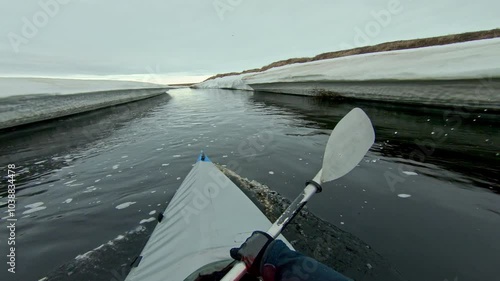 A kayaker paddles through a narrow channel in a frozen river, with snow-covered banks on either side. The sky is overcast, and a flock of birds can be seen flying overhead
