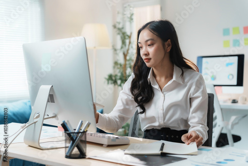 Young asian businesswoman is working on her computer in a modern office, surrounded by paperwork and digital devices