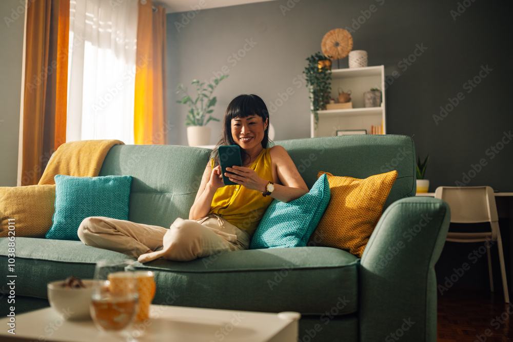 Obraz premium Asian woman lounging with smartphone on green couch, modern living room