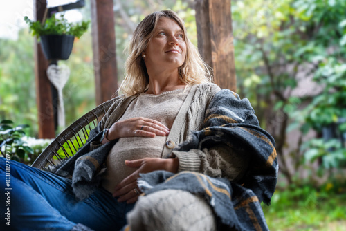 Portrait Caucasian pregnant mature relaxed woman sitting in chair on cottage terrace gently touching belly