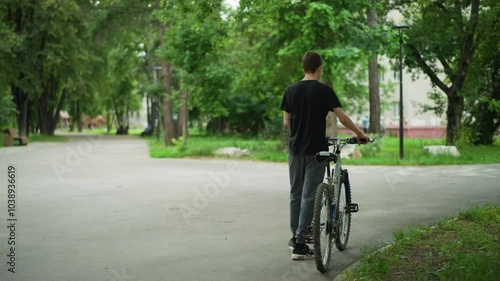 Wallpaper Mural Young boy in black top and ash pants walking alongside his bicycle in park pathway, trees and greenery line the path, while distant buildings and more trees are visible Torontodigital.ca
