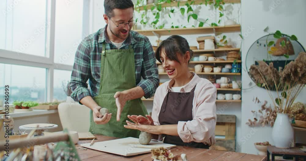 Caucasian man and woman preparing pieces of clay together. Woman sitting at table while man standing besides and showing example. Wedging clay together. Romantic activity for couples. Caucasian man and woman preparing pieces of clay together. Woman sitting at table while man standing besides and showing example. Wedging clay together. Romantic activity for couples.