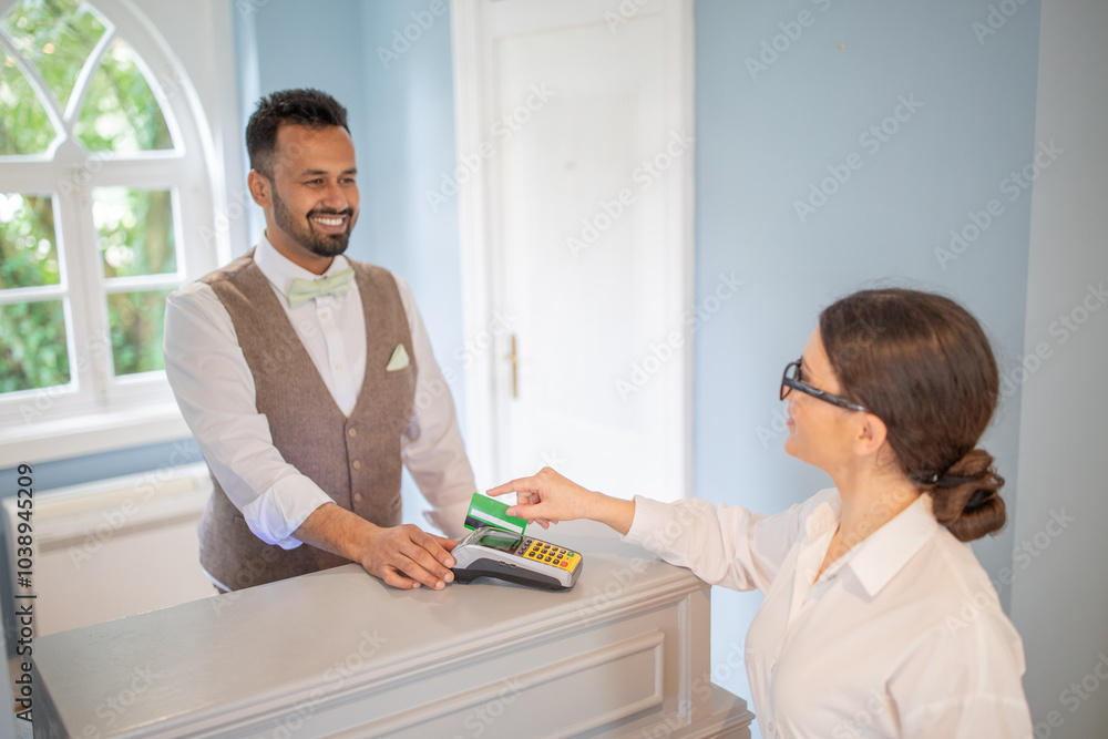 A smiling hotel clerk assists a guest during checkout in a well-lit reception area. The guest is making a payment with a card, creating a friendly interaction. A smiling hotel clerk assists a guest during checkout in a well-lit reception area. The guest is making a payment with a card, creating a friendly interaction.