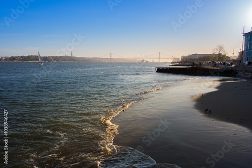 Serene coastal view at sunrise with a bridge in the distance and gentle waves lapping at the sandy shore.