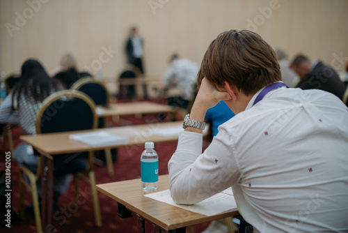 A young man is sitting at a table with a notebook on an exam. A student guy at his desk takes a test, writes a task in a notebook in the classroom