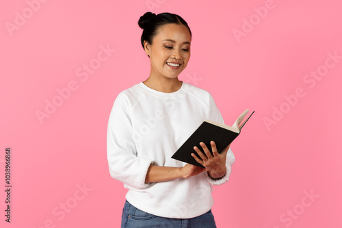Photos A young Asian woman stands joyfully reading a book, her expression vibrant and engaged