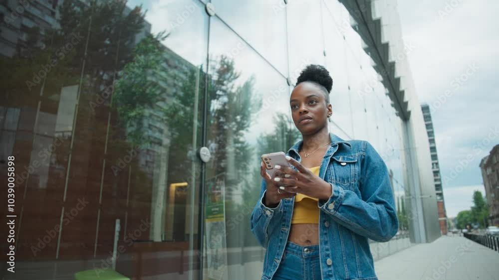 Cute African American woman walking next to modern building with tall glass walls. Using her smartphone with both hands. Searching for correct location. Checking address on mobile device. Laughing. Cute African American woman walking next to modern building with tall glass walls. Using her smartphone with both hands. Searching for correct location. Checking address on mobile device. Laughing.