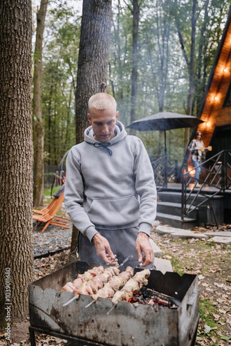 Man grilling skewers on a charcoal grill during a cozy outdoor barbecue in a forest retreat setting photo