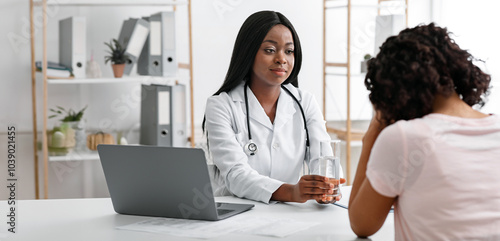 Black woman doctor comforting her upset patient, offering glass of water in clinic