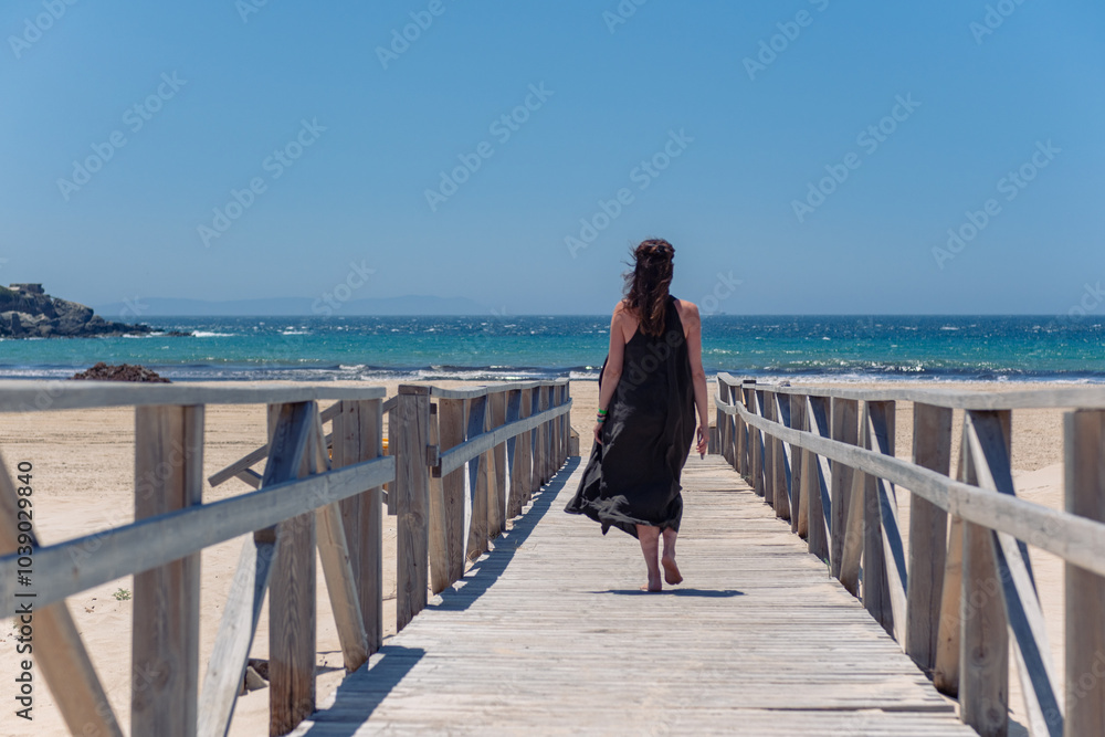 Fototapeta premium A girl in a beautiful long dress walks to the sea along a wooden bridge. Sunny day, a girl walks along a wooden bridge with railings. View from the back of a girl walking to the sea