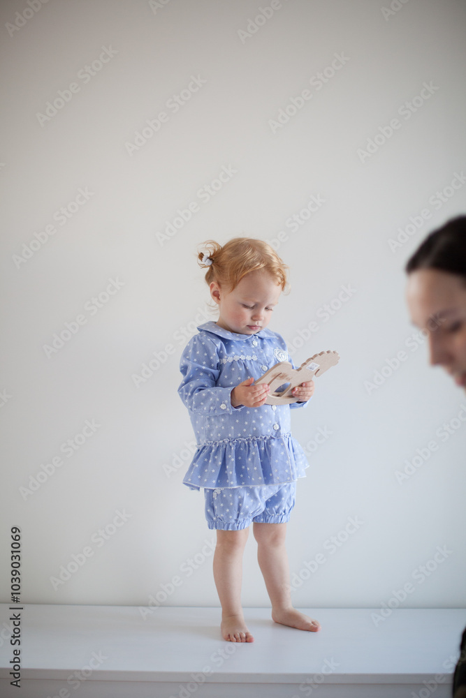 Aesthetic minimalistic portrait of Adorable beautiful smiling happy ginger-haired toddler baby girl stands on a white chest of drawers and plays with a toy horse Aesthetic minimalistic portrait of Adorable beautiful smiling happy ginger-haired toddler baby girl stands on a white chest of drawers and plays with a toy horse