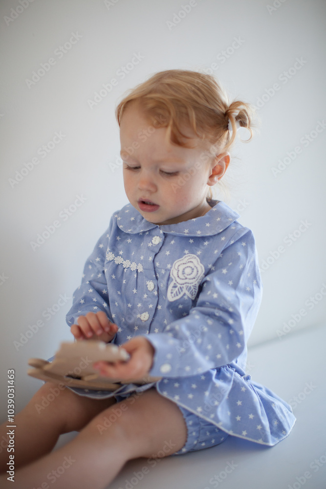 Aesthetic minimalistic portrait of Adorable beautiful smiling happy ginger-haired toddler baby girl sits on a white chest of drawers and plays with a toy horse Aesthetic minimalistic portrait of Adorable beautiful smiling happy ginger-haired toddler baby girl sits on a white chest of drawers and plays with a toy horse