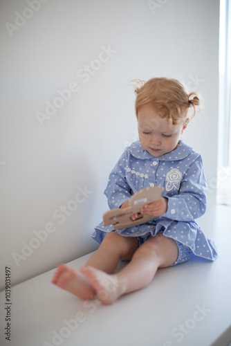 Aesthetic minimalistic portrait of Adorable beautiful smiling happy ginger-haired toddler baby girl sits on a white chest of drawers and plays with a toy horse