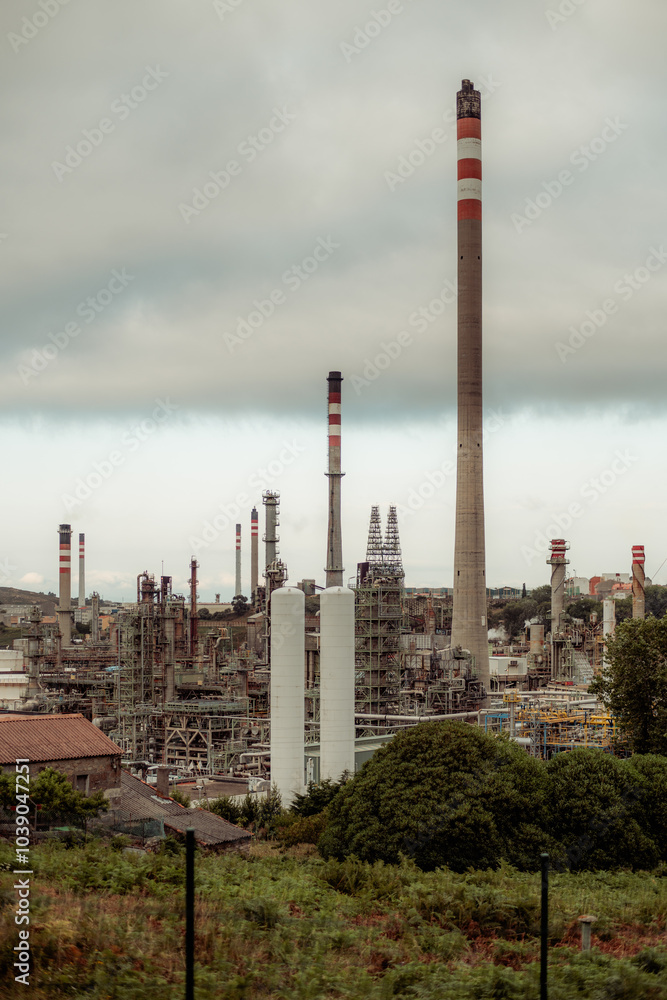 Obraz premium A vertical telephoto shot of an industrial oil refinery with multiple chimneys and cooling towers emitting fumes. Complex network of pipes, machinery, and structures, with greenery in the foreground