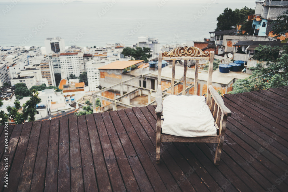 Fototapeta premium Wooden rocking chair with white cushion on a deck overlooking a hillside favela and urban buildings with an ocean view in the background. Rustic and serene setting