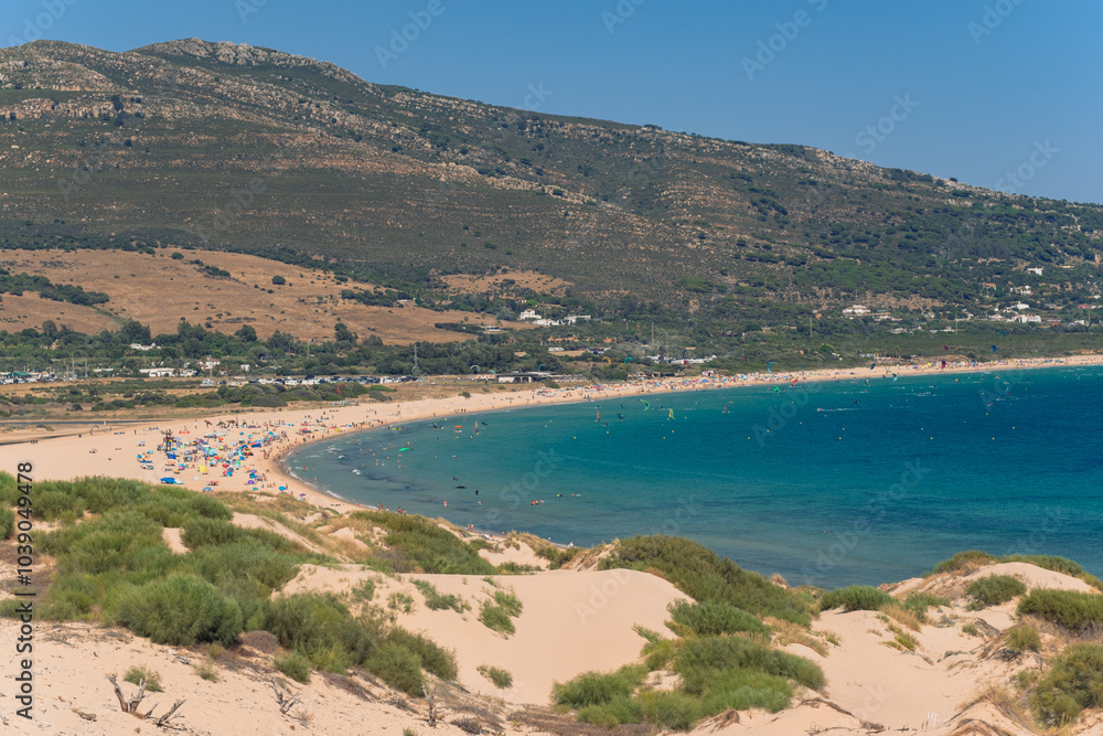 Beautiful view of a wild beach lying under a mountain. Beach with many vacationers, surfers, windsurfers. General view of a large sandy beach on the seashore in Spain Beautiful view of a wild beach lying under a mountain. Beach with many vacationers, surfers, windsurfers. General view of a large sandy beach on the seashore in Spain
