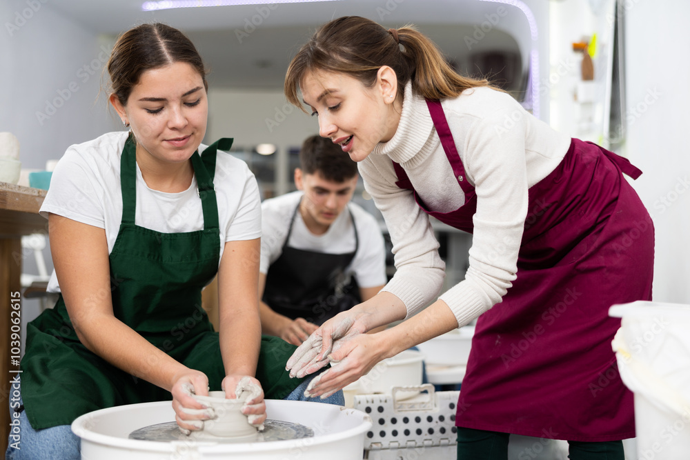 Fototapeta premium Two young women create clay product on potter's wheel in workshop