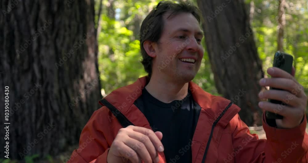 A man in an orange jacket is looking at his cell phone in the forest. A man in an orange jacket is looking at his cell phone in the forest.