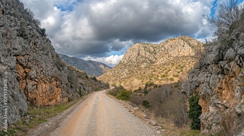 A dirt road winds through a valley, with rocky cliffs on either side. The sky is partly cloudy, with large, dark clouds overhead.