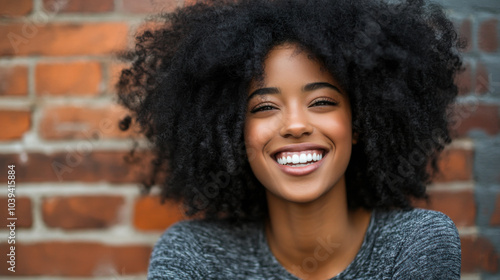 A joyful woman with curly hair smiles brightly against a brick wall in a warm, vibrant setting during the daytime