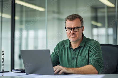  Middle-aged man with glasses working on a laptop at an office desk, blurred glass wall background, wide-angle professional photograph.