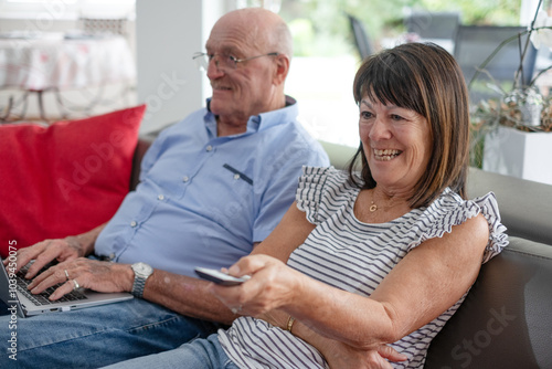 Elderly man sitting on sofa using laptop while wife watches television, two elderly pensioners relaxing in their home with large window overlooking garden