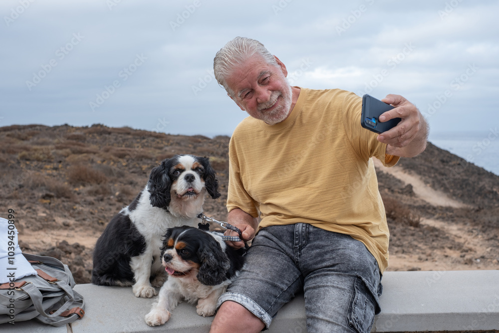 Senior smiling man sitting outdoors with his two cavalier king charles spaniel dogs along the ocean. Elderly bearded male and his best friends Senior smiling man sitting outdoors with his two cavalier king charles spaniel dogs along the ocean. Elderly bearded male and his best friends