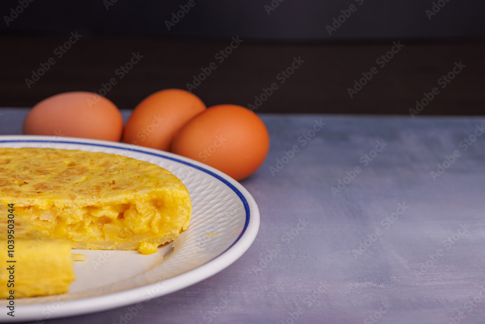 Traditional Spanish omelette with a missing piece, bread, eggs, and a red checkered napkin on a blueish concrete table Traditional Spanish omelette with a missing piece, bread, eggs, and a red checkered napkin on a blueish concrete table