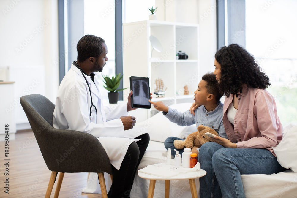 African doctor discussing x-ray results with concerned mother and young son in modern clinic. Male child showing interest in digital tablet while mother listens attentively. African doctor discussing x-ray results with concerned mother and young son in modern clinic. Male child showing interest in digital tablet while mother listens attentively.