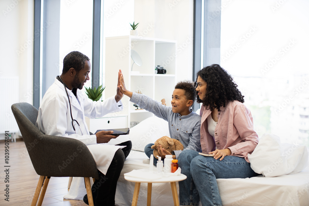 © sofiko14 - Young African boy at medical examination gives high five to African male doctor, sitting beside supportive African woman. Scene conveys positive healthcare experience, successful treatment, and trust. © sofiko14 - Young African boy at medical examination gives high five to African male doctor, sitting beside supportive African woman. Scene conveys positive healthcare experience, successful treatment, and trust.