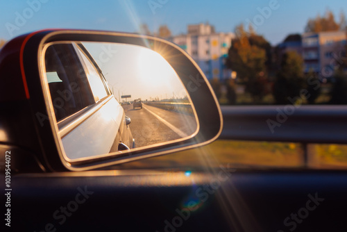 The sun is reflected in the mirror, evening road. View in the side rear view mirror of a red car driving along the highway and street
