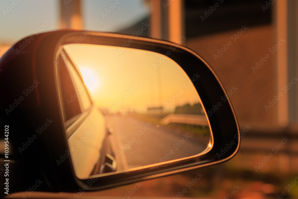 The sun is reflected in the mirror, evening road. View in the side rear view mirror of a red car driving along the highway The sun is reflected in the mirror, evening road. View in the side rear view mirror of a red car driving along the highway