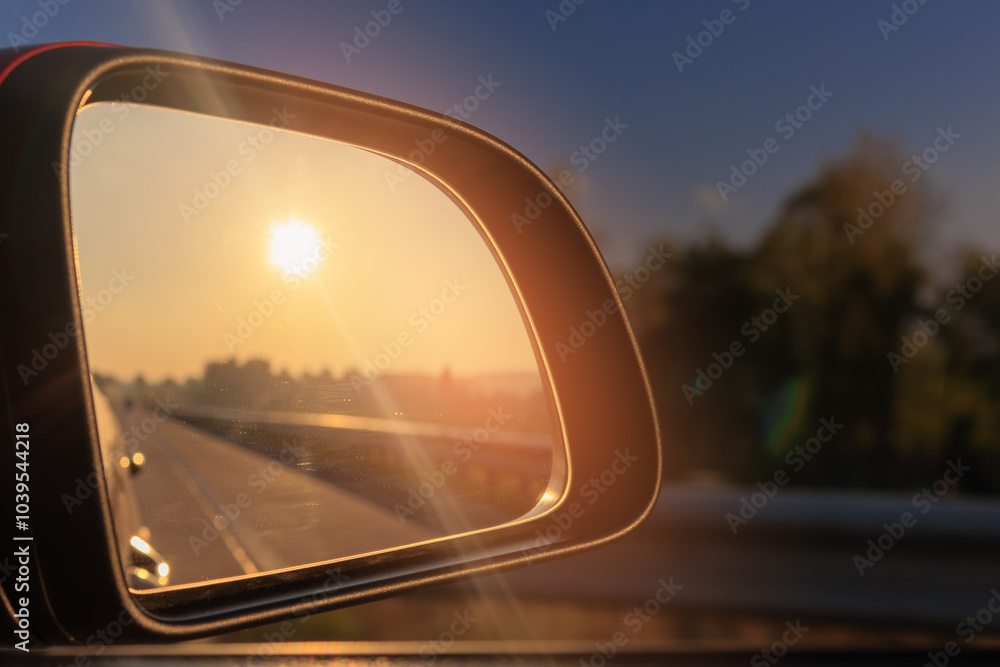 The sun is reflected in the mirror, evening road. View in the side rear view mirror of a red car driving along the highway and street The sun is reflected in the mirror, evening road. View in the side rear view mirror of a red car driving along the highway and street