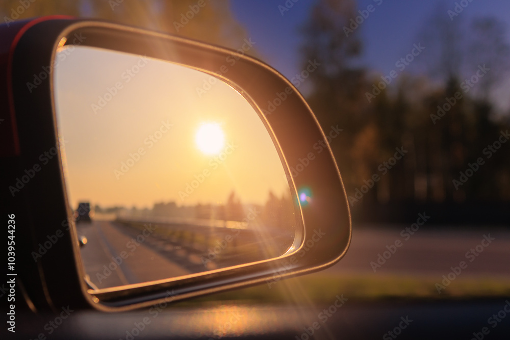 The sun is reflected in the mirror, evening road. View in the side rear view mirror of a red car driving along the highway The sun is reflected in the mirror, evening road. View in the side rear view mirror of a red car driving along the highway
