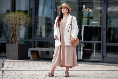 Stylish Plus size woman Standing outdoors in urban city, wearing beige dress, white blazer, wide brimmed hat, woven handbag. Model with long brown hair