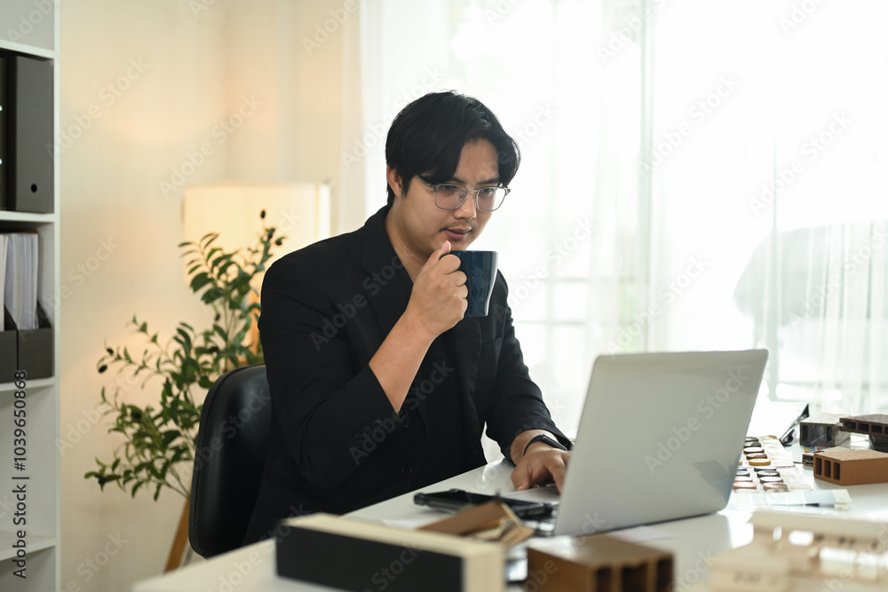 Male architect sipping coffee while working on a laptop at desk with model buildings and material samples Male architect sipping coffee while working on a laptop at desk with model buildings and material samples