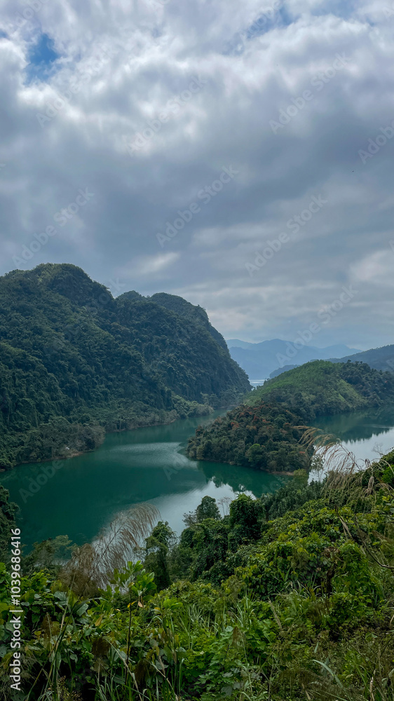 Obraz premium Serene tropical landscape with lush green mountains and a winding river under a cloudy sky, ideal for Earth Day themes