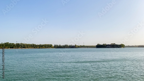 Serene waterfront view of a calm lake surrounded by lush greenery under a clear blue sky, ideal for summer relaxation