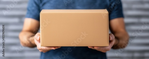 A person holds a plain brown delivery box, positioned in front of a gray brick wall, suggesting themes of shipping and receiving goods.