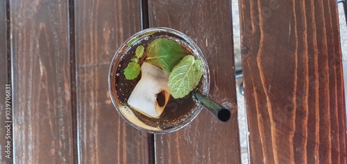refreshing drink with ice cubes, fresh mint leaves, and a black straw, served in a glass. The glass is placed on a wooden table top-down view