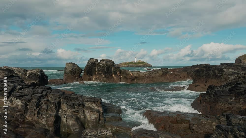 Wide shot of Cornish coast with lighthouse in the background surrounded by waves crashing on the shore. Sky is sunny with clouds. Wide shot of Cornish coast with lighthouse in the background surrounded by waves crashing on the shore. Sky is sunny with clouds.