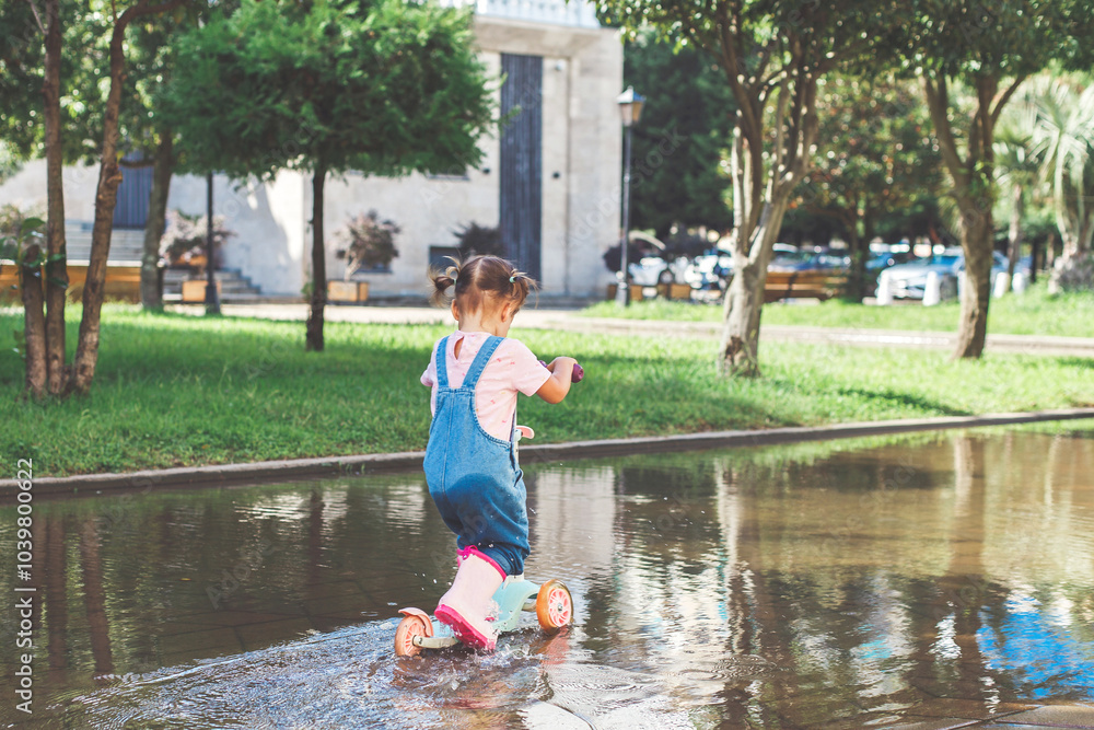 one girl in denim overalls rides a scooter through a puddle in the park, view from the back, face not visible one girl in denim overalls rides a scooter through a puddle in the park, view from the back, face not visible