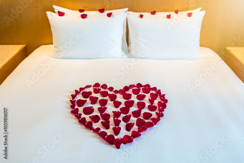 A heart shaped arrangement of red rose petals decorates a bed with white pillows
