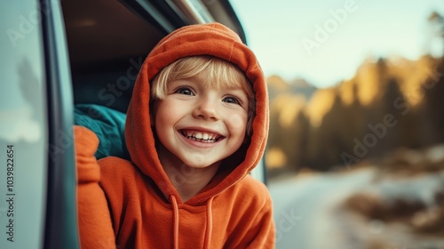 A cheerful child wearing an orange hoodie leans out of a vehicle window, smiling brightly, set against a backdrop of natural scenery with warm, golden light. photo