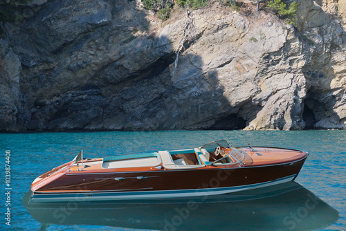 A luxury, varnished wooden boat for millionaires is anchored near a high cliff in the sea.
